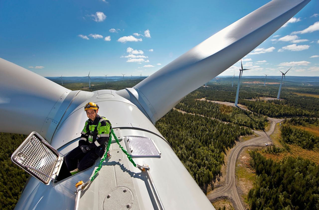 Person i varselväst och med säkerhetssele sitte på toppen av ett vindkraftverk i soligt väder.